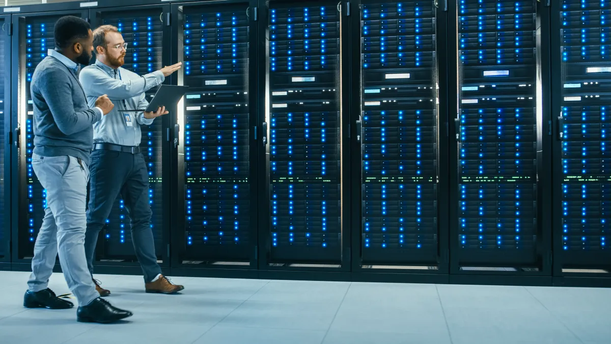 IT Technician with a Laptop Computer and Black Male Engineer Colleague are Talking in Data Center while Walking Next to Server Racks.